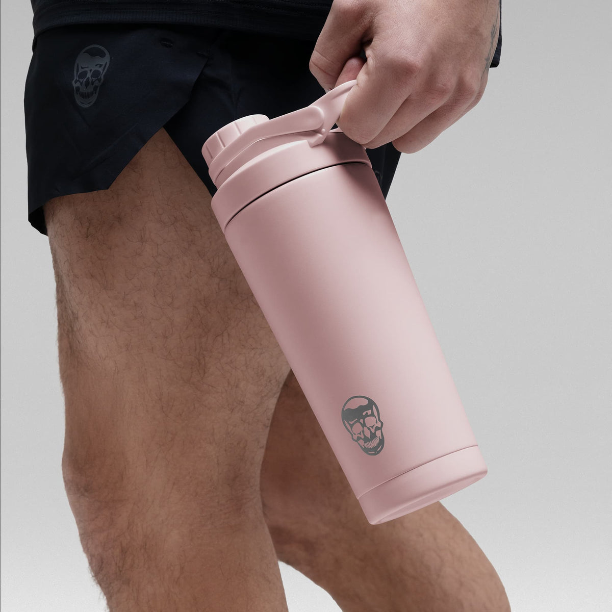 Athlete holding the Light pink stainless steel shaker bottle by the built in carry loop handle with the skull logo facing outward