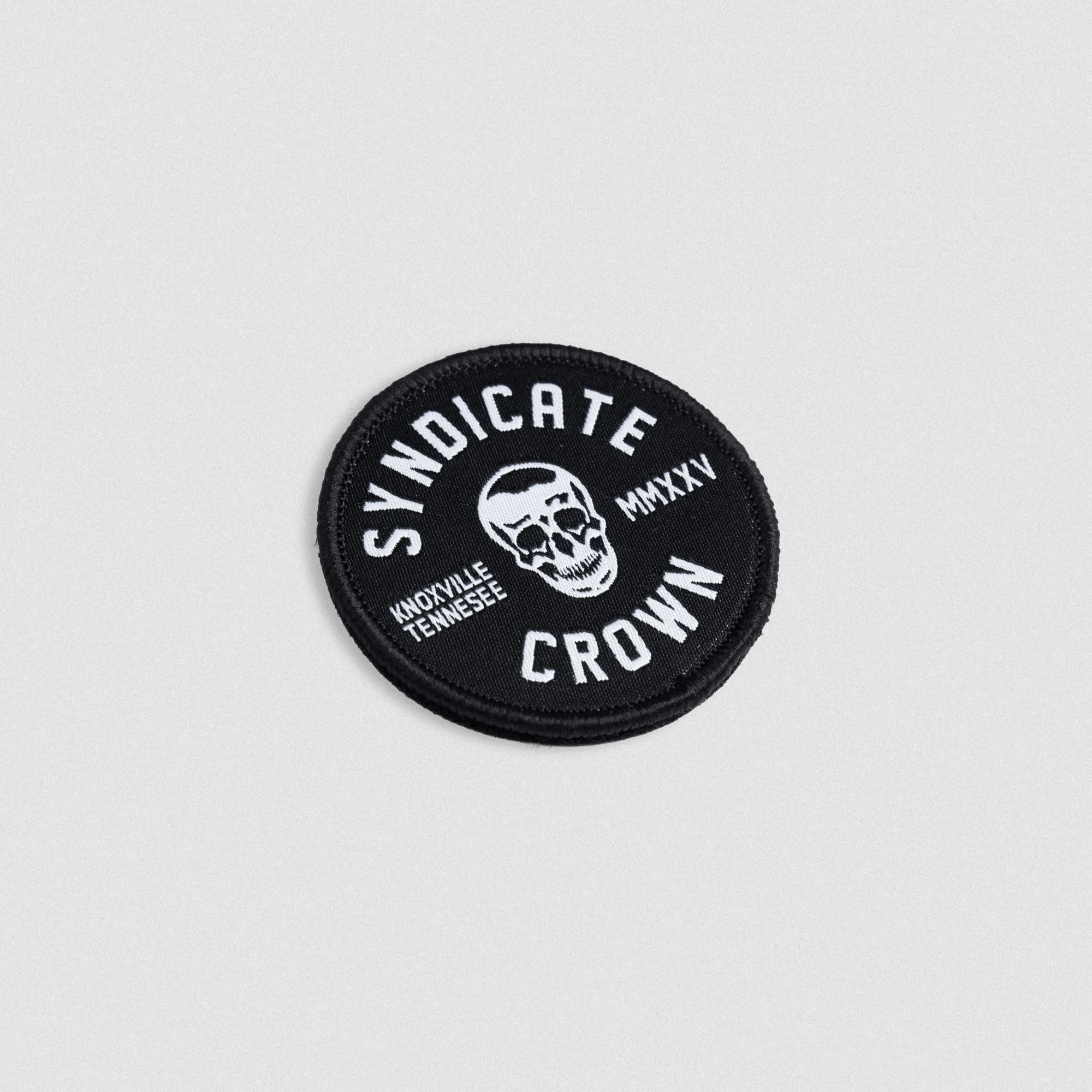 Angled view of stacked round SYNDICATE x MMXXV patches with white embroidered skull, Knoxville Tennessee text, and hook backing for attachment.
